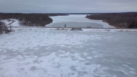 4k aerial video of a small town rural frozen lake in michigan in the winter time, small bridge, trees, on a cold overcast day