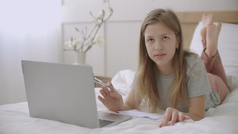 teen girl is lying on bed with laptop and exercise book and talking preparing to doing homework after school