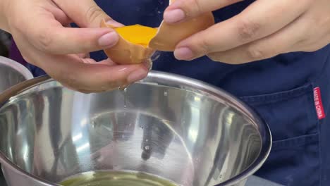 Tasty-sweet-dessert-making-in-progress,-pastry-chef-cracking-egg,-separating-egg-white-and-egg-yolk-into-different-stainless-steel-mixing-bowl,-commercial-restaurant-setting