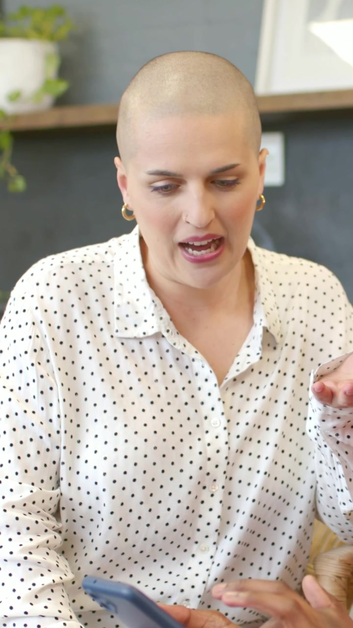 Video verticale: Donna sorridente in camicetta a pois che reagisce al telefono mostrato da una mano in un caffè con tazze rosse.