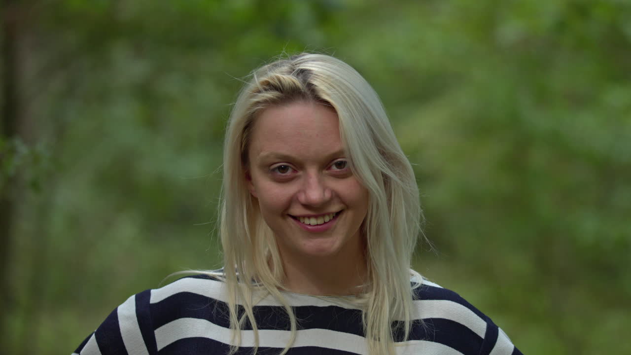 Portræt af en glad, smilende, optimistisk blond kvinde, der kigger ind i kameraet i en sommerpark eller skov.