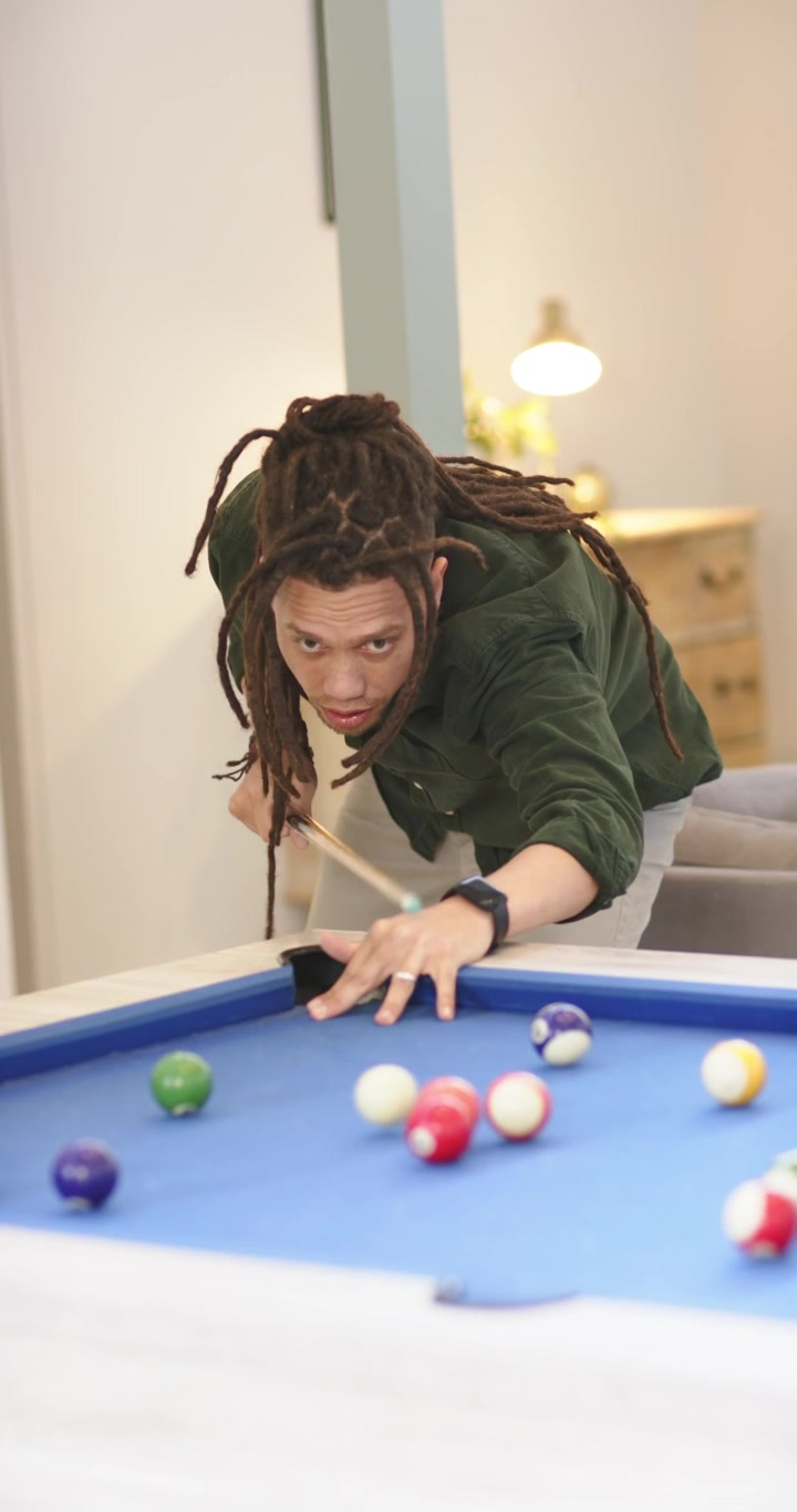 Video verticale: Uomo con i dreadlock che prepara il tiro di apertura, allineando la stecca sul tavolo da biliardo in una sala giochi e colpendo le bilie.