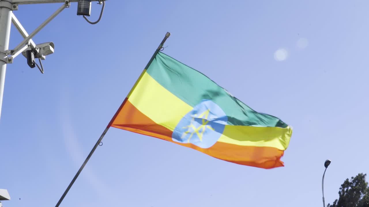 Ethiopian flag with green, yellow and red stripes and blue star disc waves on a pole under a clear blue sky, with urban CCTV cameras hinting at surveillance and security