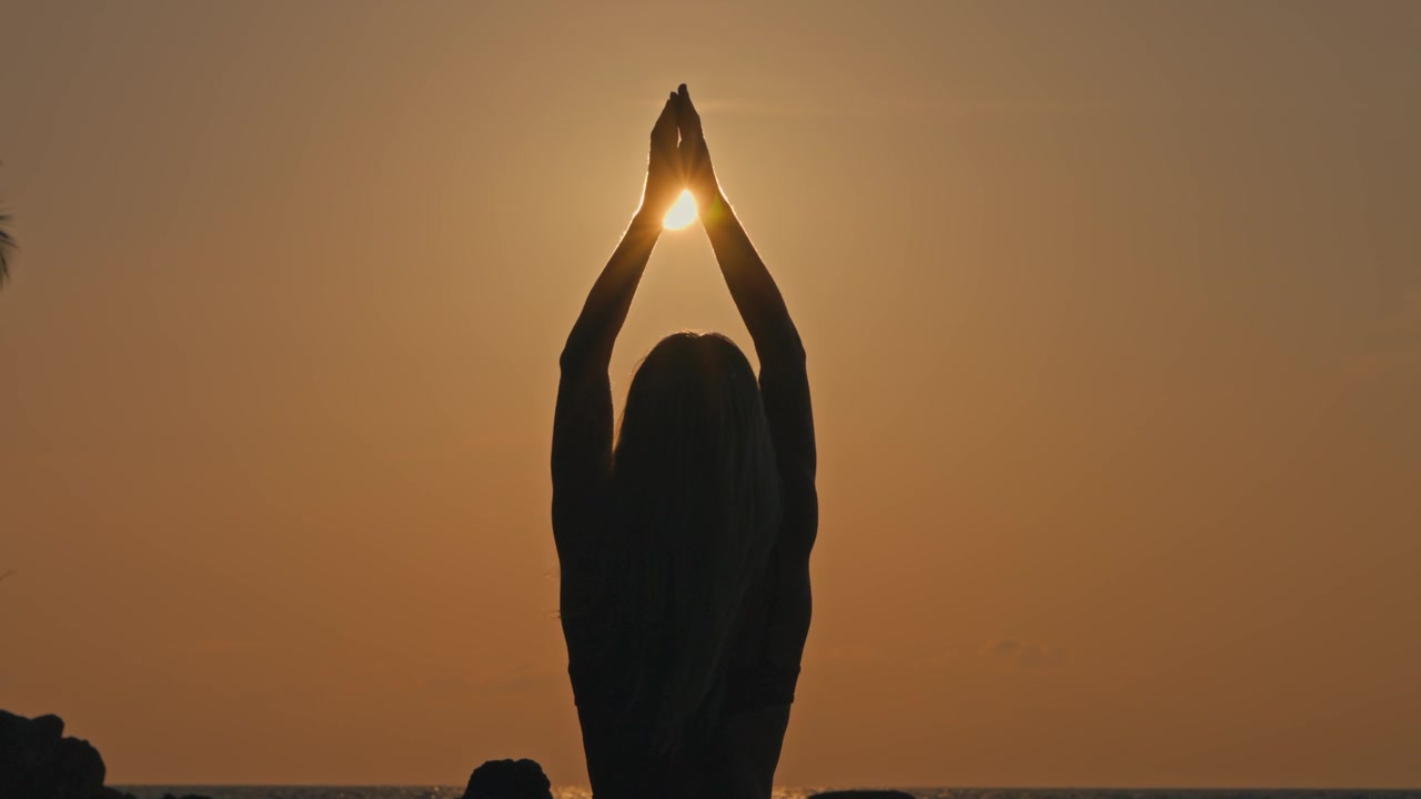 Silhouette di donna vista di spalle che esegue la posa yoga del Saluto verso l'Alto durante il tramonto contro un cielo arancione, tenendo il sole tra le mani in posizione di preghiera con le braccia alzate.