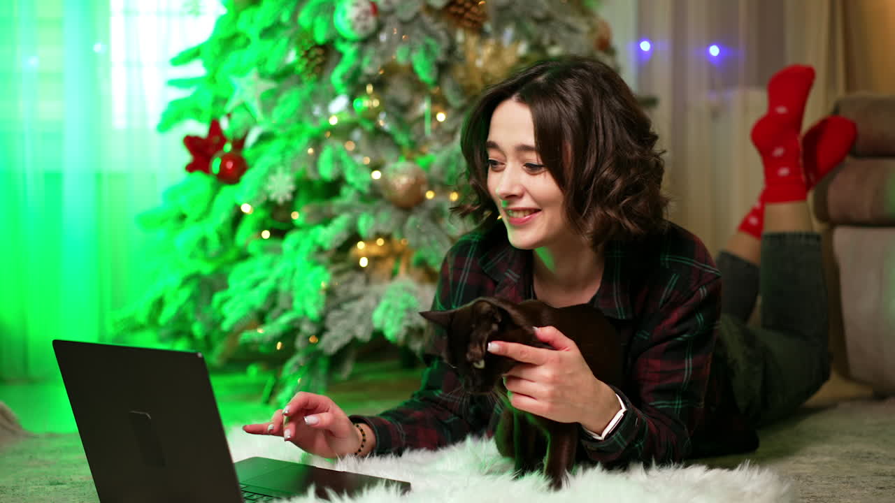 Young woman interacts with cat by laptop. A young woman lies on a cozy rug, playfully engaging with her black cat while using a laptop near a Christmas tree