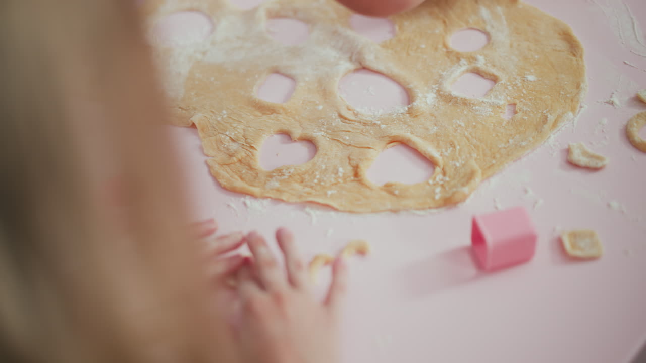 Rear view of young baker arranging rolled dough decorated with heart and circle cutouts on pink table while another child holds a cut out shape during playful baking session
