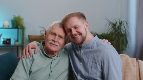 Portrait-of-mature-grandfather-with-young-adult-man-grandson-smiling-happy-embracing-hugging-at-home