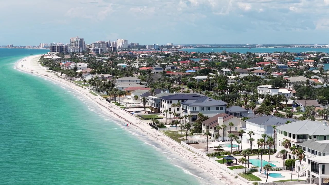 Lussuose case fronte mare con piscine costeggiano un'isola barriera di sabbia bianca accanto alle acque turchesi del Golfo con lo skyline di Clearwater Beach sullo sfondo a Clearwater Beach, Florida, USA. Ripresa aerea con drone in passaggio