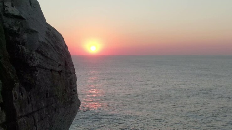 Vista aérea ao nascer do sol no mar. A câmera se move ao redor do topo de uma montanha para revelar uma vista aérea de um vasto mar contra um sol baixo.