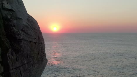 aerial view at sunrise at sea camera moves around the head of a mountain  to reveal an aerial view of a large sea against a low sun.