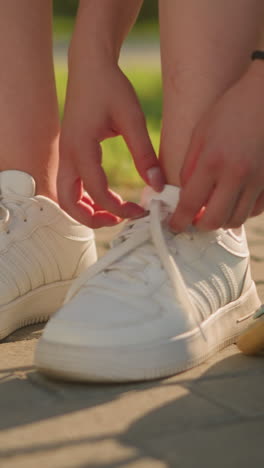 close up de la dama soltando el cordón de la zapatilla de la pierna derecha mientras estaba sentada en el pavimento, patín de rodillos acostado a su lado, sombra proyectada en el suelo con fondo de vegetación y suave luz solar