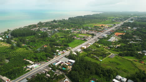 Aerial-birds-eye-shot-of-coastal-road-of-Khao-Lak-surrounded-by-green-plantation-fields-and-seascape-in-background,-Thailand
