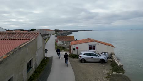 man overtaking woman in bicycle riding along the ocean promenade in île de ré island western france at village of loix, aerial follow behind shot