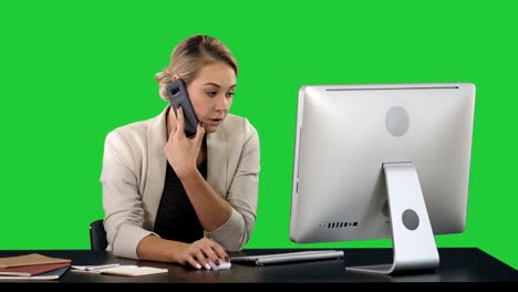 attractive young woman talking on the mobile phone and smiling while sitting at her working place in office and looking at computer on a green screen, chroma key