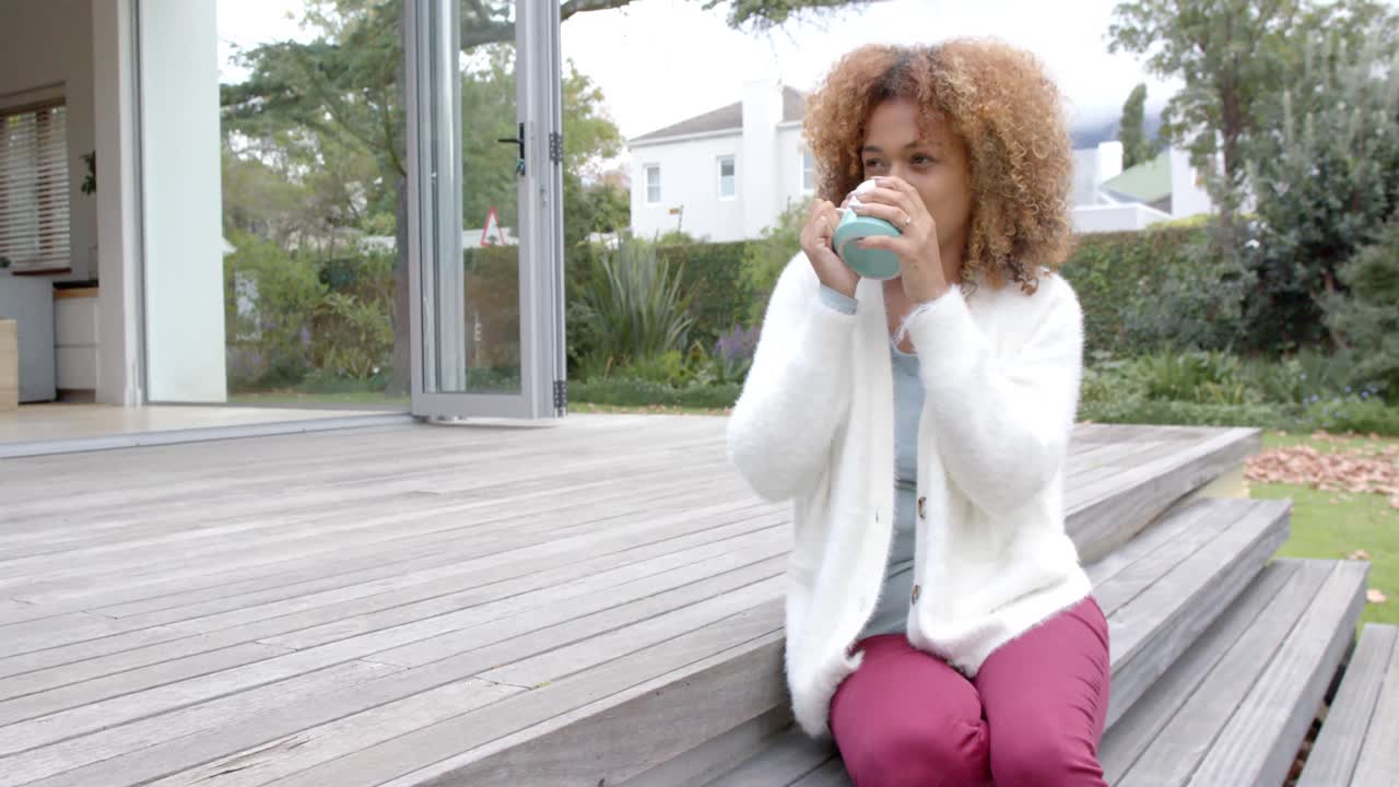feliz mujer afroamericana sentada en las escaleras y sosteniendo una taza en el jardín, cámara lenta