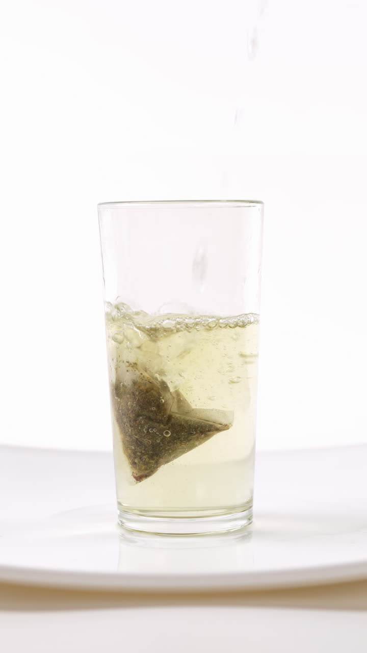 Versando acqua calda in un bicchiere con una bustina di tè, su un piatto bianco, sfondo bianco.
