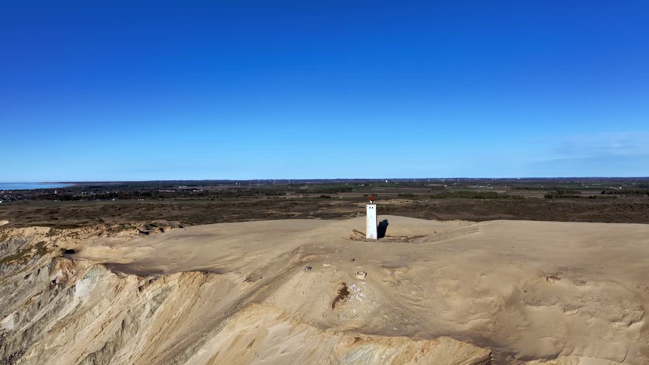 A stunning drone shot capturing Denmark's iconic stone lighthouse standing tall beside large sand dunes on the coast, showcasing the raw beauty and contrast of nature and historic architecture.
