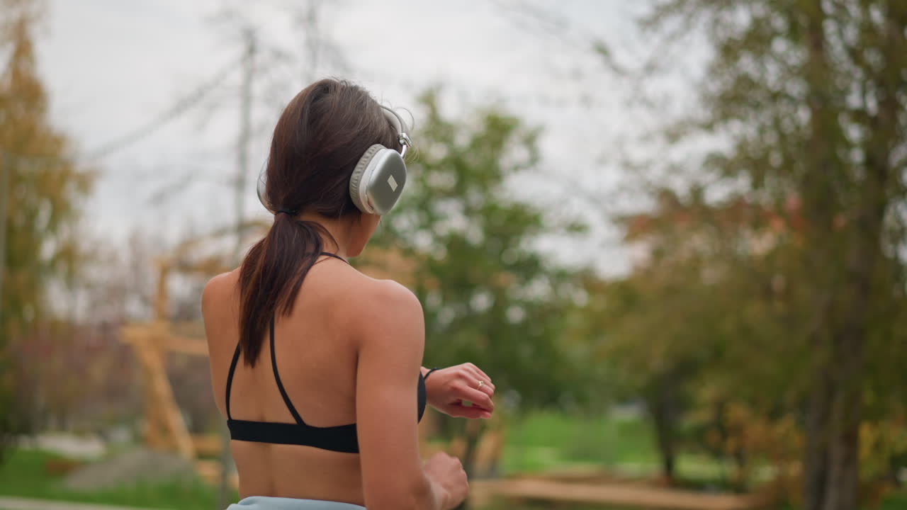 Vista lateral de una mujer joven poniéndose auriculares y caminando majestuosamente al aire libre en el parque, mostrando confianza y un estilo de vida saludable mientras disfruta de la música.