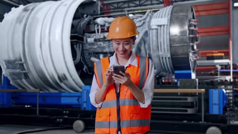 asian female engineer with safety helmet using smartphone while standing with airplane engine maintenance conducted
