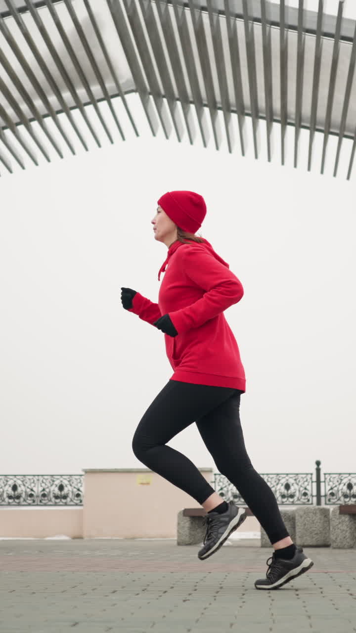 Lady in red hoodie and beanie jogging outdoors on interlocked pavement under modern pergola structure during winter in urban environment with decorative railings, benches, and snowy surroundings