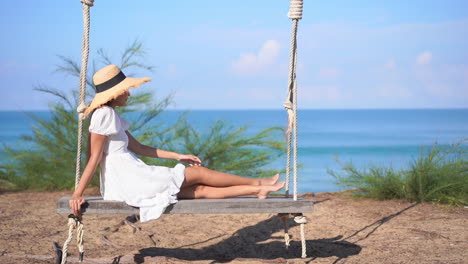 Eine-Junge-Frau-In-Einem-Sommerkleid-Und-Strohsonnenhut-Schaukelt-Sanft-Auf-Einer-übergroßen-Schaukel-Mit-Blick-Auf-Den-Ozeanhorizont