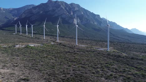 Gran-Planta-De-Energía-Eólica-Que-Está-Al-Costado-De-La-Carretera-Y-Se-Puede-Ver-Una-Gran-Montaña-Detrás-De-Ellos-Y-Un-Cielo-Muy-Despejado-Con-Pasto-Verde-En-El-Suelo.