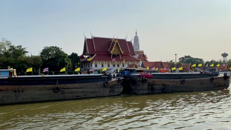 traditional thai temple by river, barge in foreground, clear sky, late afternoon, cultural landscape