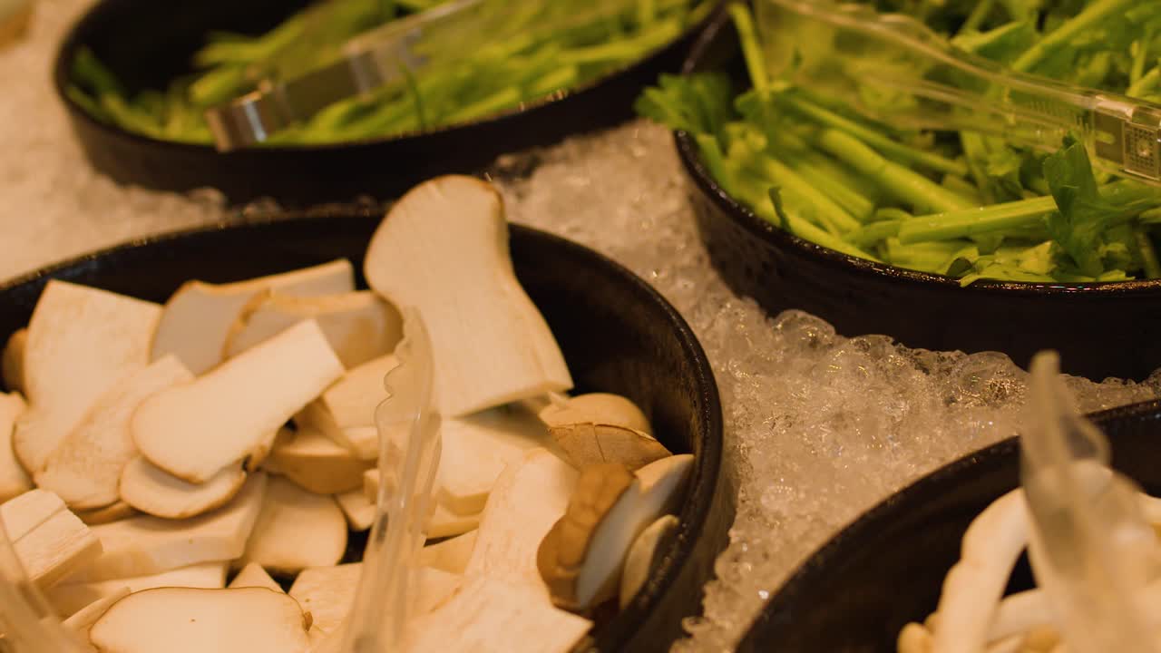 Assorted fresh mushrooms, greens, and corn displayed on ice under warm lighting at buffet bar