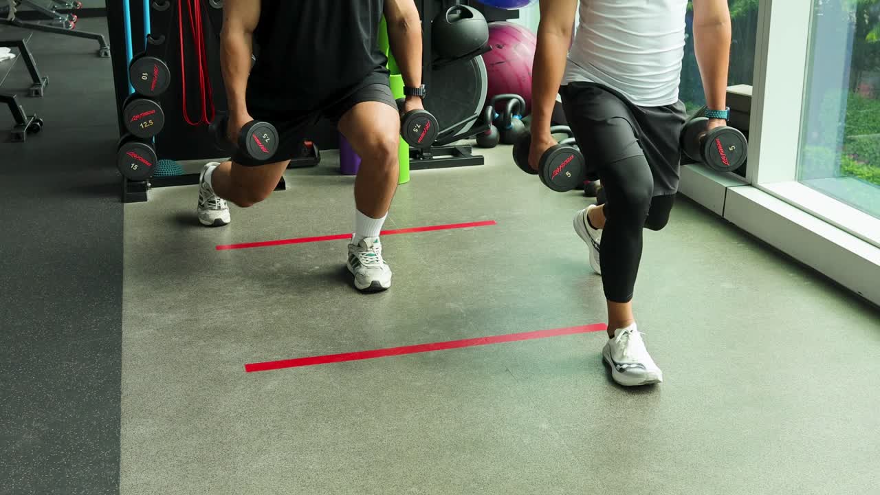 Two men perform synchronized dumbbell lunges in a sunlit modern gym, demonstrating controlled movement