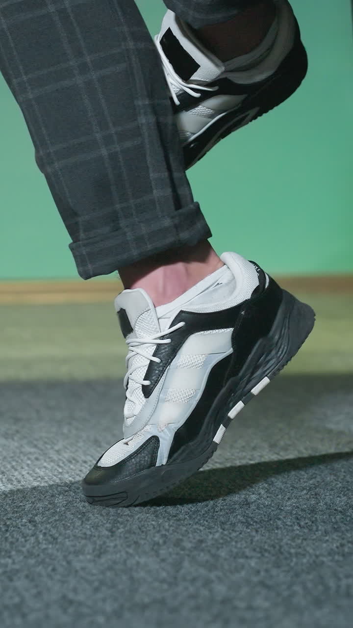 toma de primer plano de una pierna masculina con zapatillas de deporte elegantes blancas y negras con pantalones a cuadros grises. pie ligeramente levantado del piso de alfombra gris, declaración de moda moderna y casual