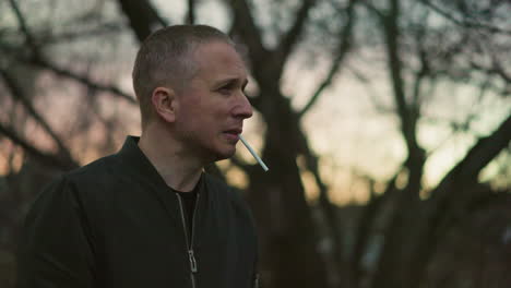 a man in a green jacket stands outdoors at dusk, holding a cigarette to his mouth, deep in thought, with blur view of cars moving from behind