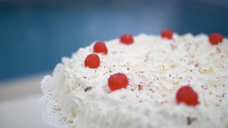 Delicioso-Pastel-De-Crema-De-Cumpleaños-Blanco-Con-Chispas-Y-Detalles-De-Cerezas-De-Caramelo-Rojo-En-Un-Jardín-Con-Una-Piscina-En-El-Fondo-En-Cámara-Lenta