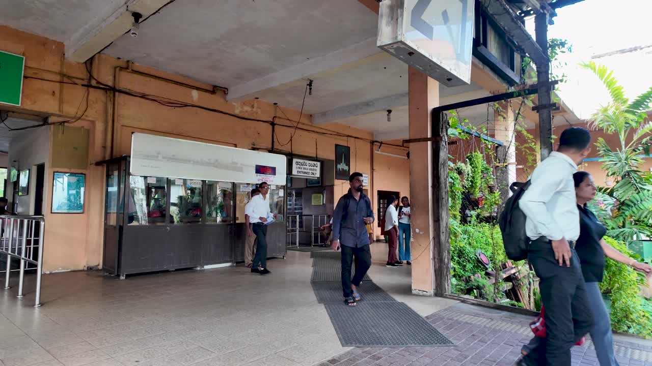 People enter the Kandy train station platform, showcasing daily commuting in Sri Lanka. The scene captures travelers amidst lush greenery and a bustling atmosphere typical of public transportation.