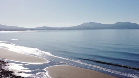 Aerial-view-rising-above-shimmering-Newborough-beach,-Anglesey-with-Snowdonia-mountain-range-landscape