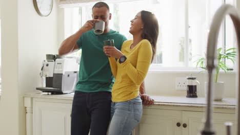 happy biracial couple standing in kitchen, drinking coffee and laughing