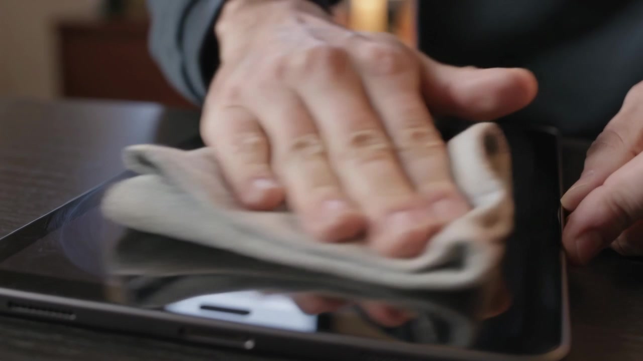 Hænder lægger en foldet klud og tørrer tabletglasset af på et mørkt bord for at fjerne pletter, iført sweater.
