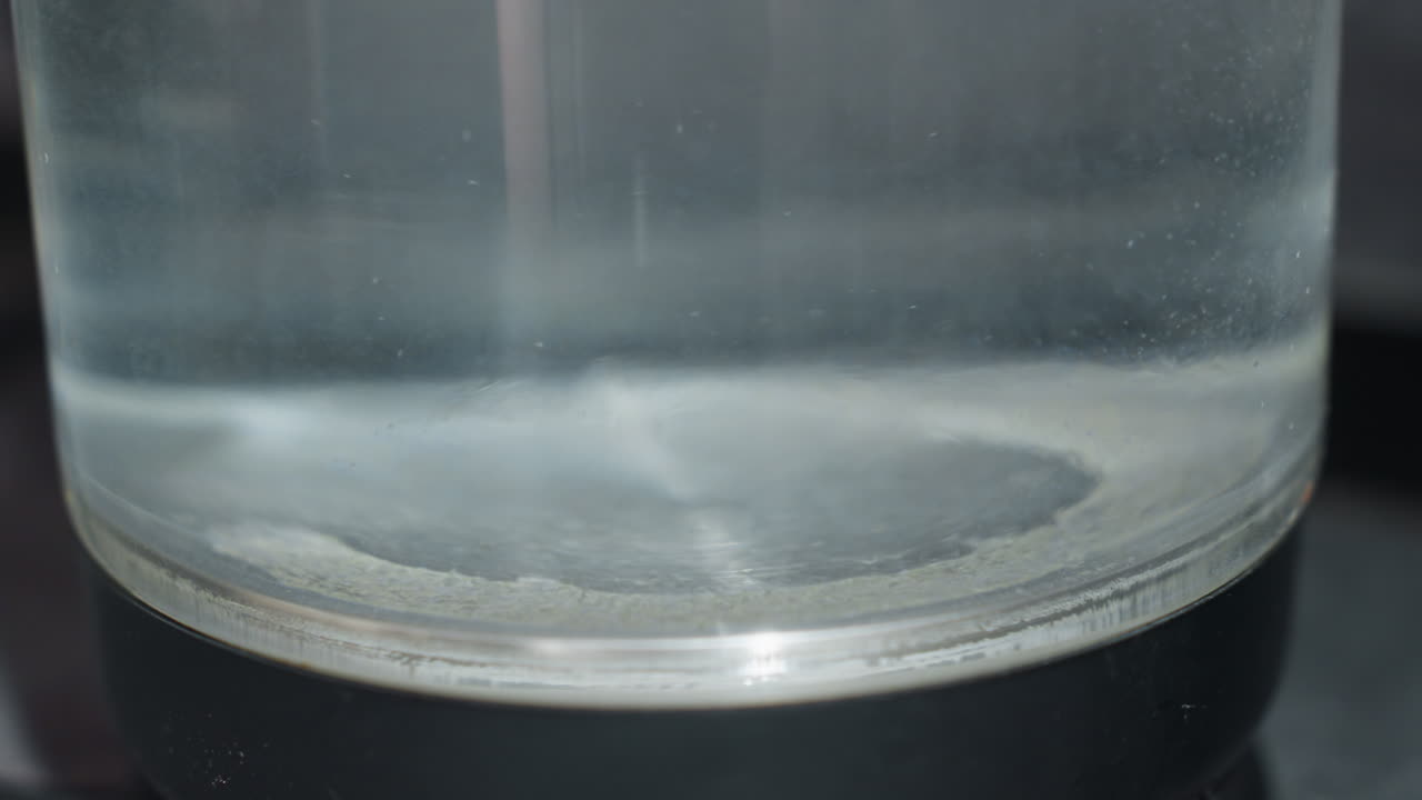CloseUp Glass Beaker Bottom Showing White Precipitate Ring And Slow Swirl, Clear Solution With Faint Blue Reflection, Clinical Bench Setting, Scientist Monitoring Reaction And Measurement