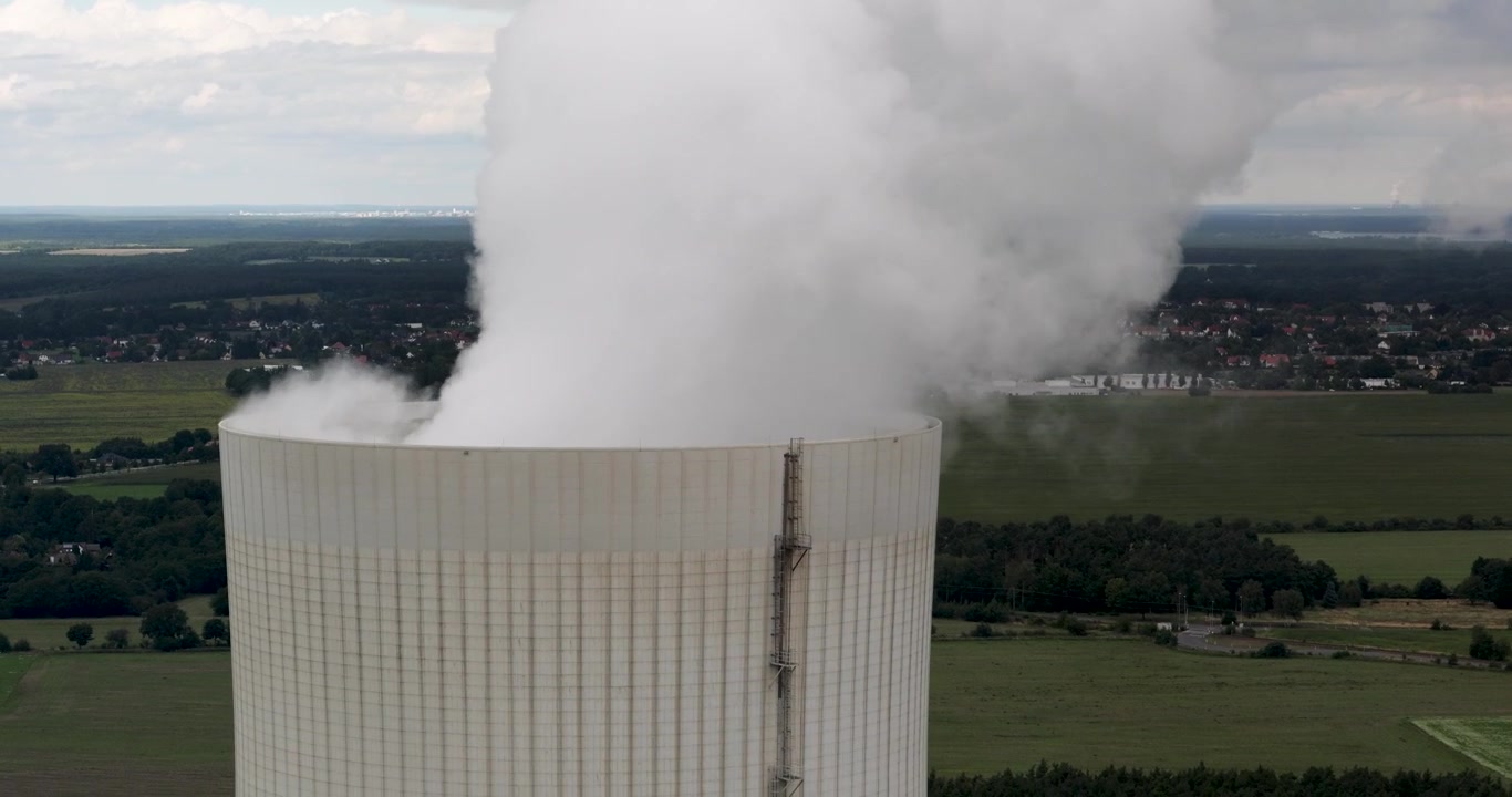 Vista dettagliata di una torre di raffreddamento di una centrale elettrica con una scala di servizio, che emette fumo bianco nel cielo, affacciata su città rurali nel contesto dell'eliminazione graduale del carbone in Germania (Kohleausstieg)