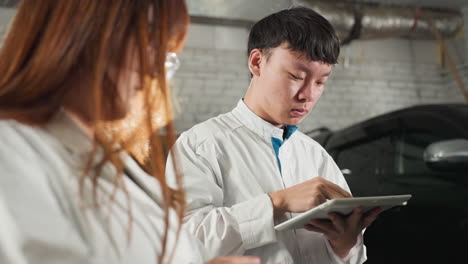 students in lab coats take notes on tablets while observing a mechanic demonstrating tool usage on an engine, industrial automotive workshop filled with equipment, and mechanical parts in background