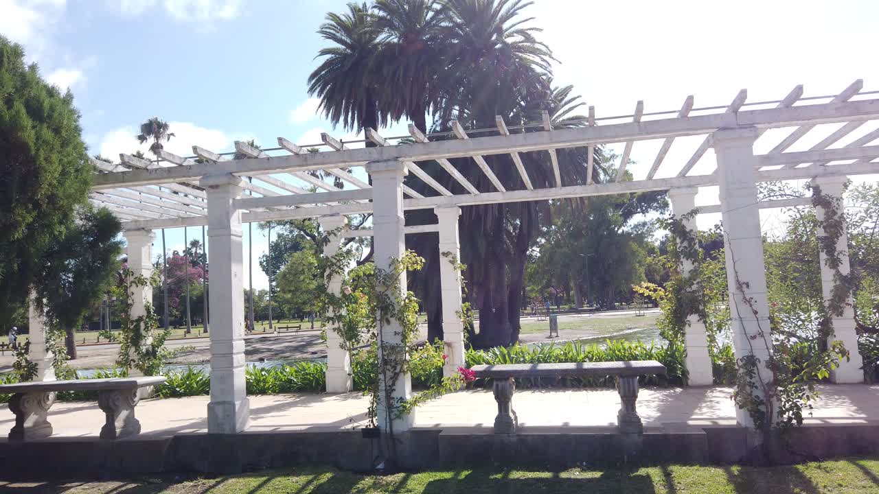 Schwenkaufnahme der Landschaft des Rosengartens von Palermo, Buenos Aires, Argentinien, mit einer weißen griechischen Pergola und joggenden Menschen im Hintergrund.