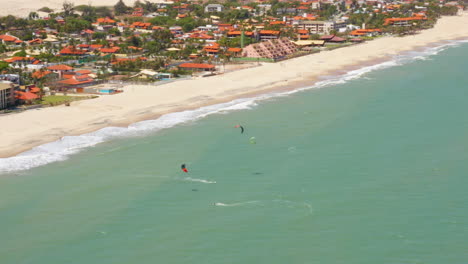 Vista-Aérea-De-Personas-Practicando-Kitesurf-Y-Un-Pequeño-Pueblo-Alrededor,-Cumbuco,-Ceará,-Brasil.