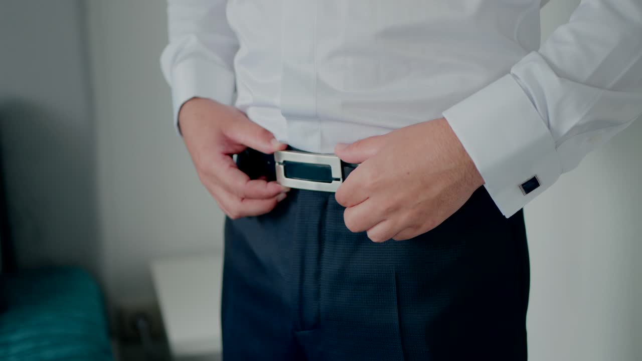 man adjusting silver belt buckle on dark trousers with white shirt and cufflinks near blue chair indoors