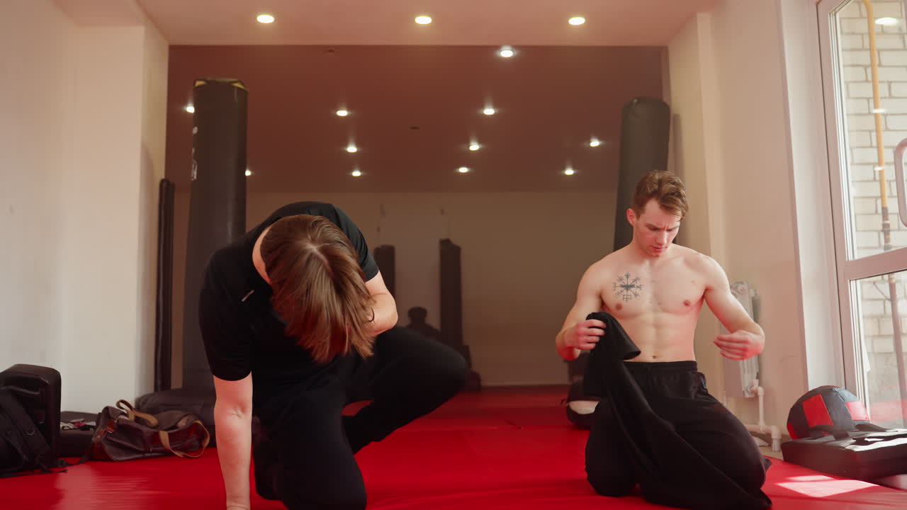 Two men inside gym walking on red mat after workout, removing their shirt while other holds gear, training area with punching bags, sports bags, natural light through window, fitness lifestyle