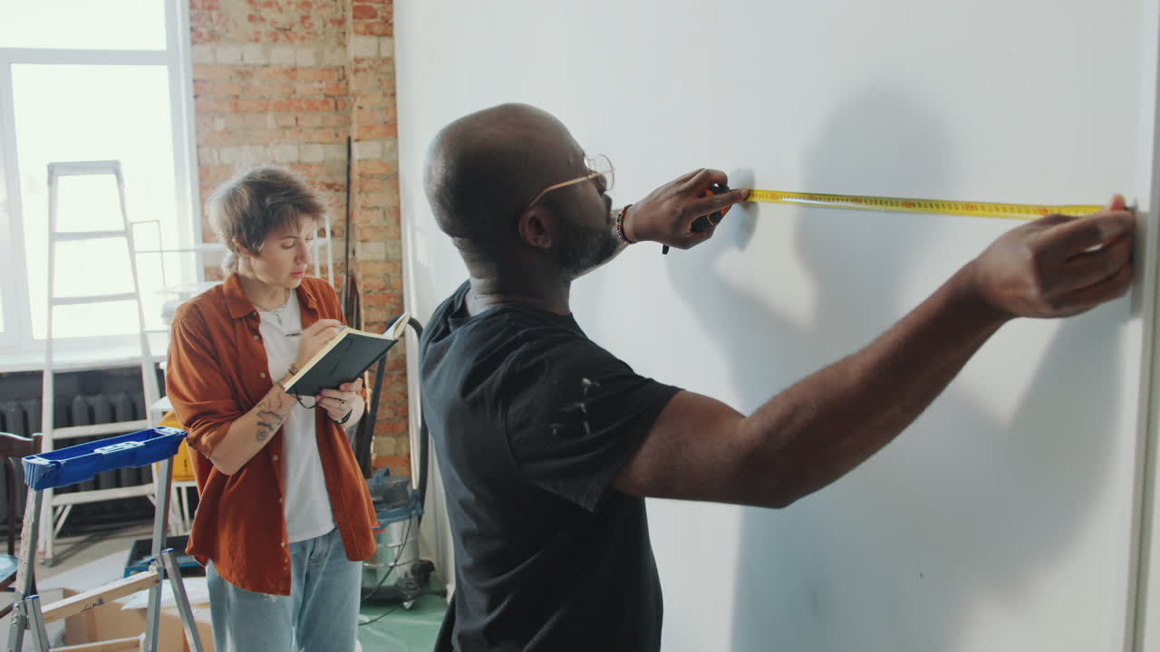Man Taking Wall Measurements Telling Wife Writing Notes during Home Renovation