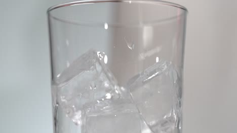 ice cubes in tall clear glass having clear liquid poured on from metal jigger close up slow mo 4k