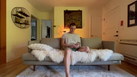 Caucasian-male-sits-on-fur-covered-couch-reading-a-book-in-their-apartment