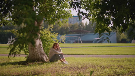 woman sits thoughtfully on grassy ground under tree, resting her hand on her leg, gazing downward in peaceful contemplation, background includes greenery, poles, and stadium