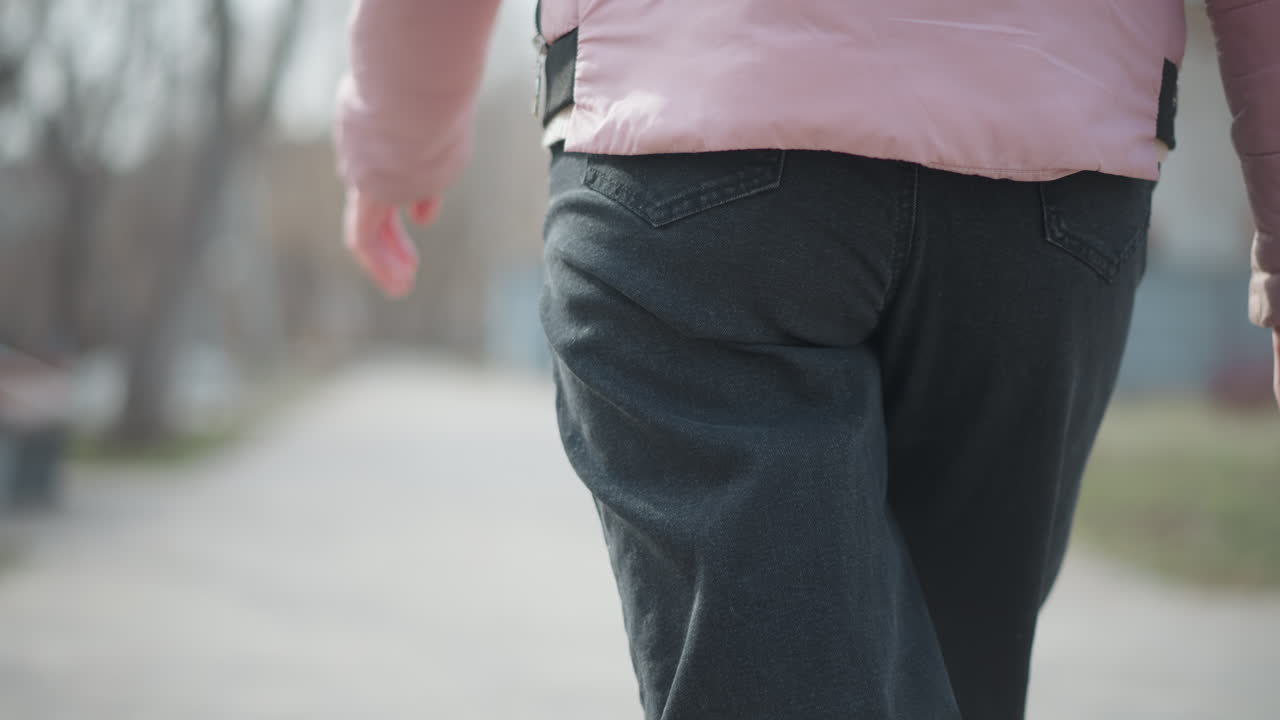 Close-up rear view of woman walking outdoors during winter in pink padded jacket and black jeans, arm swinging gently, with soft natural light and blurred background of trees and park walkway path