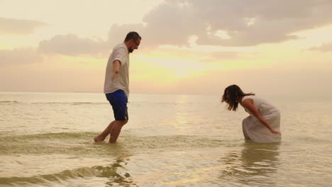 Pareja-Joven-Divirtiéndose-En-El-Mar-Espolvorear-Agua-El-Uno-Al-Otro-Al-Atardecer