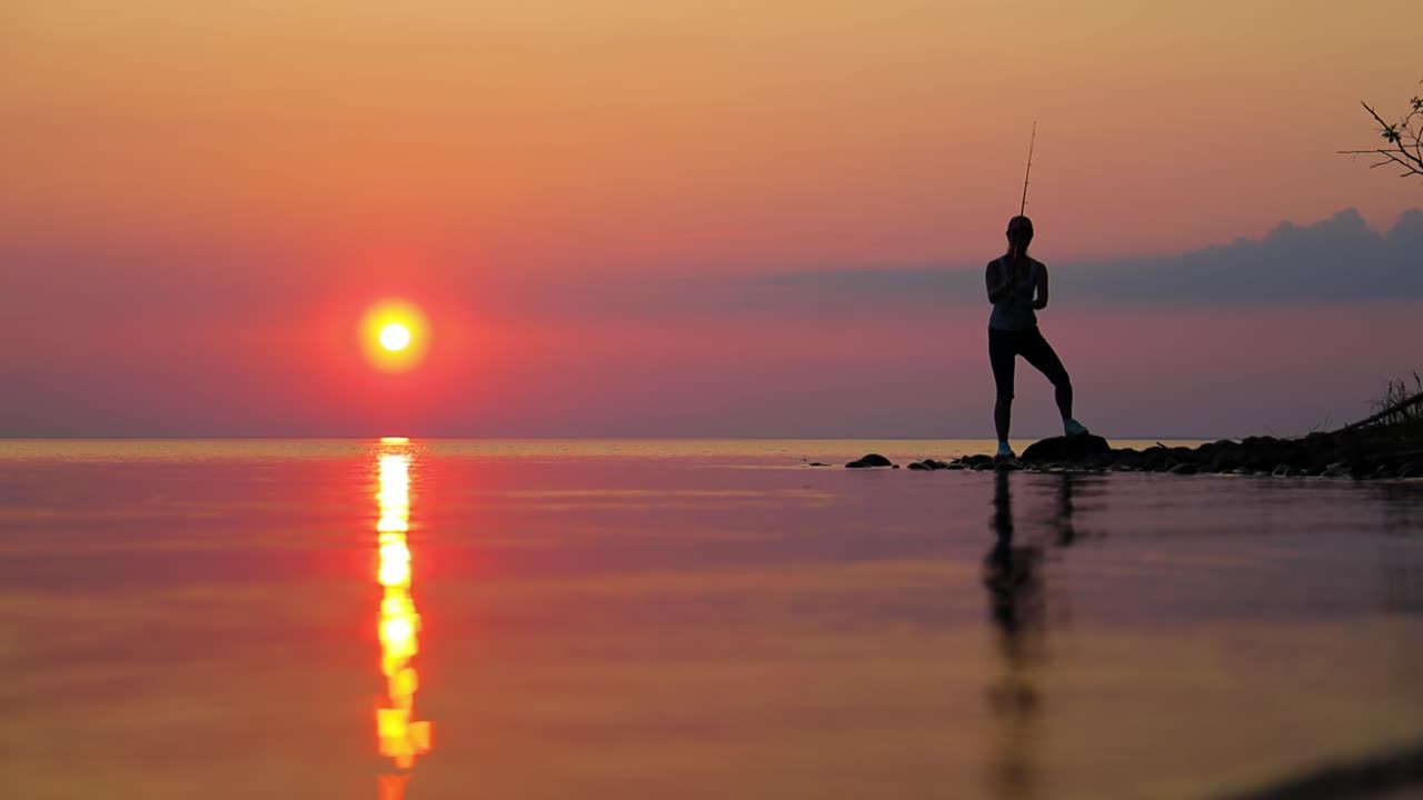 Wanita memancing dengan joran pancing berputar dengan latar belakang matahari terbenam.